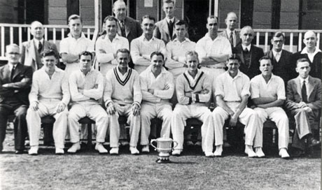 Photograph of the twelve players of Horden Cricket Team, accompanied by nine other men, posed in front of the pavilion; a trophy can be seen on the floor in front of the players