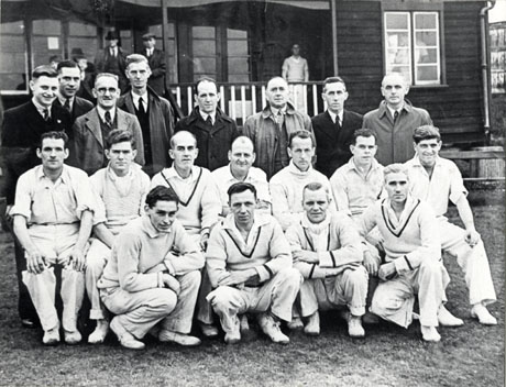 Photograph of eleven players in cricket whites, posed with eight other men, in front of a pavilion, on the verandah of which five other men can be seen; the photograph has been described as Horden County Cricket Team