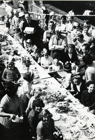 Photograph, taken from above, of a long table running from the bottom right to the top left of the photograph, laden with plates of food and flowers; children are sitting at the table and adults are standing behind them watching them and handing them food and pouring tea; the occasion for the street party has been identified as the Coronation of Queen Elizabeth II and its location as Meadow Avenue, Horden