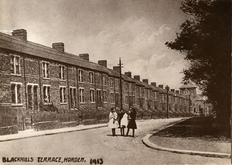 Photograph showing the exterior of Blackhills Terrace, Horden; the terrace runs from left to right the length of the photograph with the church in the distance at the extreme right of the picture; three girls carrying dolls are standing in front of the terrace in the middle distance