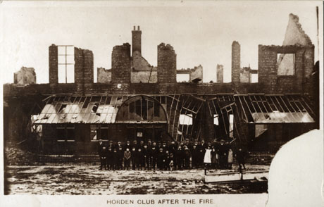 Photograph of the exterior of a building that has suffered a fire; above the ground floor only part of the walls and only one window remain; on the ground floor part of a damaged glass roof can be seen and light through the damaged walls; a group of approximately thirty six adults and children are posed outside the building; the photograph is entitled Horden Club After The Fire