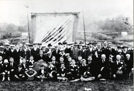 Photograph of a group of approximately fifty five adults and children grouped in front of a banner in the open air; both the banner and the countryside behind it are indistinct; the children on the front row of the group are holding musical instruments and a large drum can be seen; the photograph is identified as Horden Junior Band, 1928