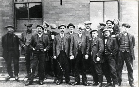 Photograph of a group of fourteen men standing in front of the wall of a brick building in which there are two windows through one of which a man is looking; one of the group is holding a child; all of the group, who are dressed in suits, may be coal miners; the photograph is described as Group outside Royal Hotel, 3rd Street, Horden