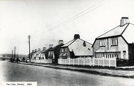 Postcard photograph entitled The Villas, Horden. 8493 showing the surface of the road and, beyond it, the fronts of semi- detached houses with bay windows and mansard roofs; in front of the houses are small gardens with light-coloured fences; a few indistinct figures can be seen on the pavement; the houses have been identified as Wraith Terrace