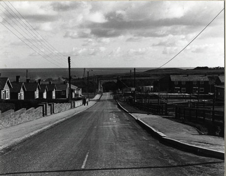 Photograph of a road leading away from the camera towards a railway tunnel and beyond that the sea; on the left hand side of the road single-storeyed buildings can be seen; on the right-hand the playground of a school can be seen; the photograph has been identified as Road Leading To Railway Bridge And Cowan's Farm (Horden)