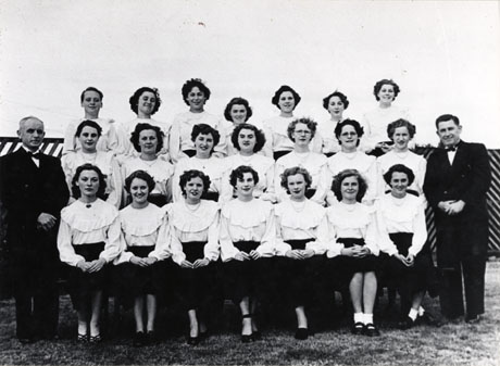 Photograph of the twenty one female members of the choir of Thorpe Street Methodist Church, Horden, posed in the open air in front of a fence; they are accompanied by two men in evening dress; the members of the choir are wearing uniform dark skirts and elaborate blouses