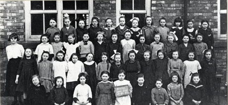 Photograph of forty seven girls aged approximately twelve years grouped outside a brick building with a woman, presumably their teacher; a girl on the front row is holding a notice which reads: Horden Senior Girls. Standard 4A 1921