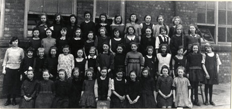 Photograph of forty seven girls aged between approximately fourteen and eleven years, posed outside a brick building with a woman, presumably their teacher; a girl on the front row is holding a notice reading 1924; the photograph has been identified as Horden Senior Girls