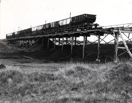 Photograph of fifteen railway trucks on a metal bridge over rough grass, with coal waste beneath the bridge; the photograph has been identified as Teeming by Horden Colliery, 1976
