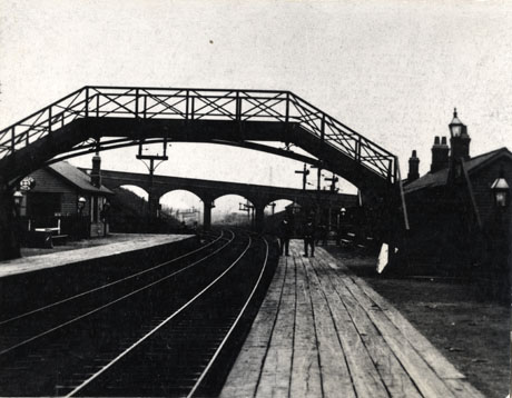 Photograph of a railway station showing the line receding away from the camera under a footbridge and road bridge; both platforms, and buildings on each platform, can be seen; two indistinct men can be seen on the right-hand platform; the station has been identified as Horden Station