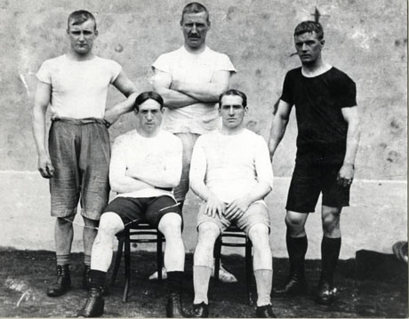 Photograph of three men standing behind two men sitting on chairs; all five men are wearing singlets and shorts; the photograph has been identified as The Two Mordue Boys versus The Three Stone Brothers 1914 Horden Hand Ball Alley