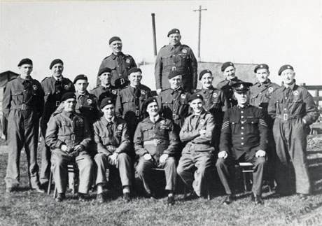 Photograph of a group of sixteen members of the Royal Observer Corps with a sergeant in the police force; the men are posed outside with part of an unidentified building behind them; all the men are in uniform