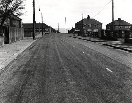 Photograph showing a road receding from the camera towards the sea in the distance; on the left of the photograph a fence and the top storey of houses can be seen; on the right in the distance semi-detached houses on a road leading into the first road can be seen; the photograph has been identified as Windsor Terrace and Alder Road 1976