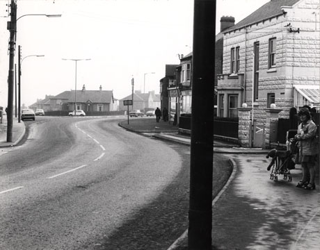 Photograph showing a road leading to the right; a road joins the first road from the left, and at the junction a bungalow and three parked cars can be seen; on the right of the first road a detached house and shops can be seen; a woman with a push chair can be seen on the pavement to the right of the photograph