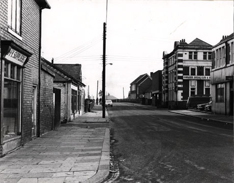 Photograph looking along a road with the facades of shops on the left and the Comrades Social Club and the facade of a school in the distance on the right; a car is parked in the road in the distance; the road has been identified as Windsor Terrace, Horden