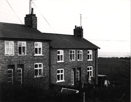 Photograph of the exterior of four terraced houses each with three windows and a door; in the distance to the right the sea can be seen; in front of the houses are small gardens; they have been identified as Railway Cottages, Beach Road 1976