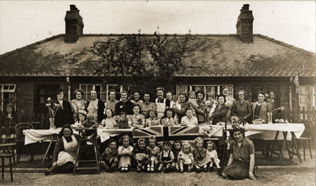 Photograph showing a trestle table, laid with a table cloth and a Union Jack, in front of two semi-detached bungalows; two women and nine children are sitting on the ground in front of the table; nine children are sitting behind the table on which plates of cakes and sandwiches and vases of flowers can be seen; behind the children sitting at the table, fifteen women are standing, three of whom are holding infants; the photograph has been identified as Street Party, (Adam and Eve Houses, Horden); the street party was most likely held to commemorate the coronation of Queen Elizabeth II