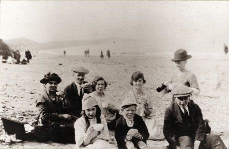 Photograph showing a group of people on a beach with indistinct figures and indistinct shoreline and hills in the distance; the group consists of a middle-aged man and woman, three girls aged between ten and eighteen years and three boys aged between seven and fifteen years; two girls are drinking from tea cups and the man , woman, a boy and a girl are eating sandwiches; one boy is pouring liquid from a Thermos flask; the photograph is described as Sands at Horden 1925 Bennett Family