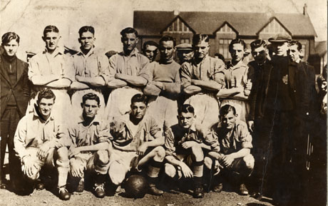Photograph of the eleven members of the Horden Juniors Football Team, accompanied by eight other men; the photograph is being taken with a half-timbered building in the distance