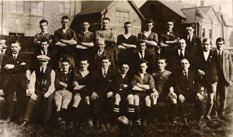 Photograph of the twelve members of the Athletic Football Team Horden Colliery, accompanied by eleven other men; the group is posed before the rear of a large building