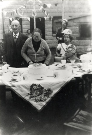 Photograph showing a table in the open air covered in an elaborately embroidered table cloth and laid with plates and cups, which appear to have been used; behind the table is an elderly woman cutting a large cake which is also on the table; beside her is an elderly man and a small boy and a small girl wearing fancy hats; behind them two women, the facade of a house and bunting can be seen; the occasion of the street party has been unidentified but it is thought to be the Coronation of Queen Elizabeth II