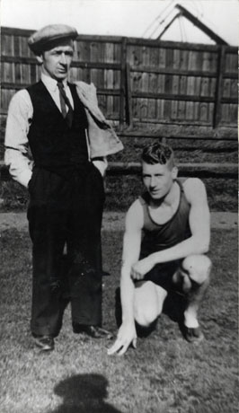 Photograph of a man wearing shorts and running vest kneeling on the ground, accompanied by a man dressed in trousers, waistcoat, tie and cap, standing beside him in front of a high fence; the two men have been identified as J. Bestford, footrunner, and J. Hamilton, coach