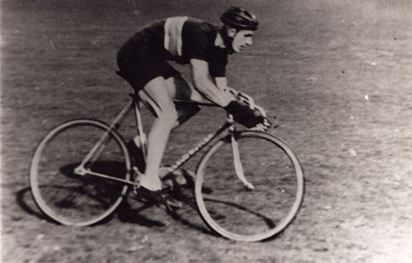 Photograph, close-up, of a man wearing racing clothes and helmet on a bicycle; the man is photographed from the side and has been identified as Harry Harland, bicycle champion 1940s