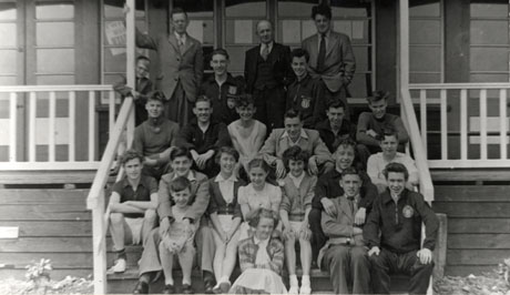 Photograph twenty two men, women and children sitting on the steps of the verandah of a woden building, presumably a pavilion; three men at the back of the group are middle aged and dressed in suits; the rest of the members of the group are young and some are dressed in sporting clothing; the group has been identified as Horden Sports Club Members and Officials