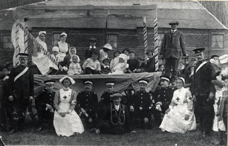 Photograph of a carnival float in front of a house; on the float are eleven children, a woman, and two women dressed as nurses; in front of the float are seven men in the uniform of the Saint John Ambulance Brigade and two nurses; in front of these people is a man wearing the insignia if the Good Templars; a man is standing on the shafts of the float; people can be seen indistinctly surrounding the float; it has been identified as St. John Ambulance Carnival Float, Horden, 1917