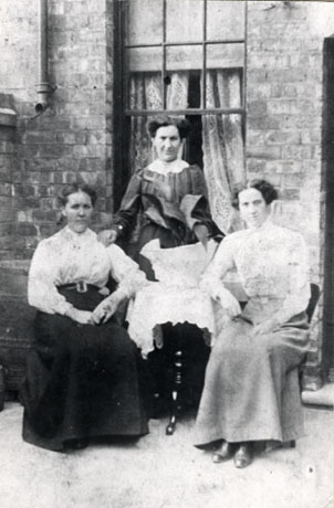 Photograph of two women sitting resting their arms on a small table and a third woman standing behind the table in the back yard of a house; the wall of the house and a window in the wall and part of the wall dividing the yard from that next door can be seen; the women are dressed formally in elaborate blouses and long skirts; a large pot plant can be seen on the table; the women are identified as Lizzy Byers and her sister, 1918