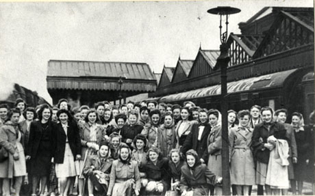 Photograph of a group of approximately sixty young women grouped on a railway station; the roofs of the station can be seen, as can the tops of two locomotives on the lines on either side of the platform where the women are grouped; the women are described as Horden and District Girls who worked in the Aycliffe Munitions Factory, 1939 - 1945