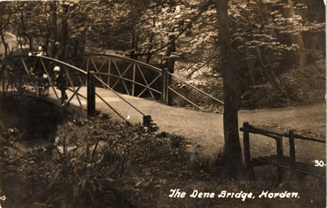 Postcard photograph entitled The Dene Bridge, Horden, showing the end of a bridge with open steel railings on either side of the road of the bridge; the bridge is surrounded by dense trees; two very indistinct figures can be seen on the bridge; the photograph has been described as The Dene Bridge, Castle Eden, leading to Hesleden from Horden, 1930s-1940s