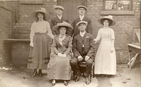 Photograph of a group of three women and two men in the backyard of a house; a face can be seen at the window of the house; two men and two women are standing round a man and a woman who are sitting on chairs; woman sitting down is wearing an elaborate hat and skirt and jacket with large buttons on them; the seated man is wearing a suit and button hole and cap; the women standing are also wearing elaborate hats and clothing; what is possibly a poss stick can be seen at the right of the photograph; the photograph has been identified as recording a wedding at Horden