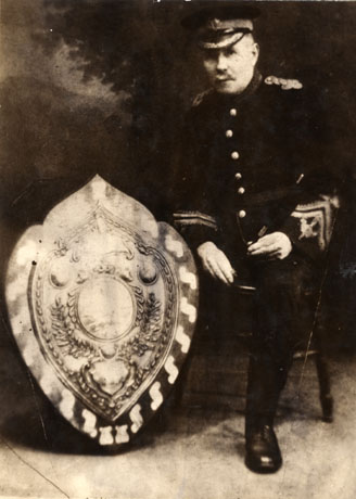 Photograph of a middle-aged man in uniform sitting on a chair with a large trophy on the floor next to him; he is holding a baton in his hand and the photograph appears to have been taken in a photographer's studio; he has been identified as Joe Foster, Bandmaster