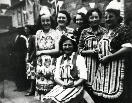 Photograph of six women wearing pinafores and fancy hats with indistinct houses and two indistinct figures behind them; three of the women are wearing identical striped pinafores; the photograph has been identified as Morpeth Street Coronation Party, Horden, 1953