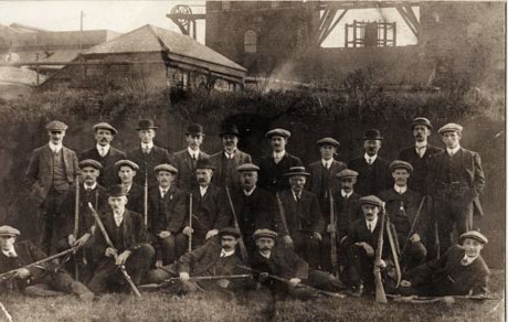 Photograph showing twenty four men posed in front of the buildings at Horden Colliery; all the men are dressed in suits and ties and are holding rifles; the photograph has been identified as Horden Officials' Rifle Club