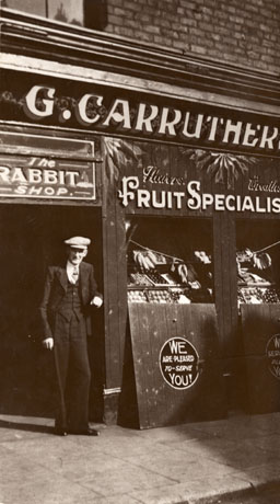 Photograph showing a man dressed in suit and tie and cap standing in the doorway of a shop with the words G. Carruthers Fruit Specialist Flowers Wreaths above the shop window; the displays of fruit and vegetables in the shop window can be seen; above the doorway are the words The Rabbit Shop; the man has been identified as G. Carruthers