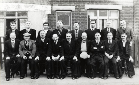Photograph of seventeen elderly and middle-aged men posed outside a house, the door and window of which can be seen; all the men are dressed in suits, but are in the main bare-headed; they have been identified as members of the Horden Comrades' Club