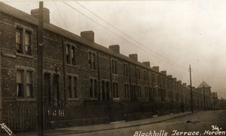 Postcard photograph entitled Blackhills Terrace, Horden, produced by Lawson. Pegswood.; the photograph shows the exterior of a terrace of houses running the length of the street, with the roof of a church at the end of the street