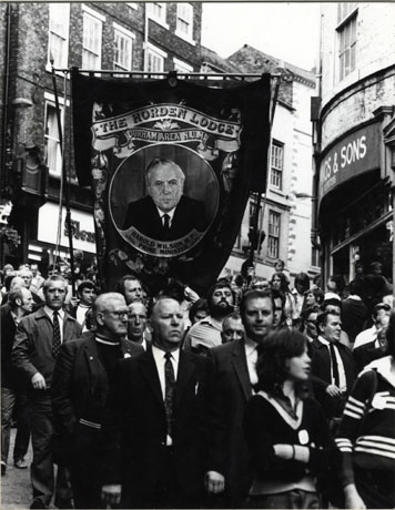 Photograph of the Horden Lodge banner being carried down the top of Elvet Bridge in the City of Durham; the banner is surrounded by a crowd of people and features the portrait of the head and shoulders of Harold Wilson, Labour Prime Minister of Great Britain, 1964-1970 and 1974-1976