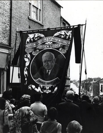 Photograph of the Horden Lodge banner being carried down the top of Elvet Bridge in the City of Durham; the camera is looking down Elvet Bridge, and Old Elvet can be seen in the distance crowded by people attending the Gala; the banner is surrounded by people with their backs to the camera; the banner carries a portrait of Emmanuel Shinwell, prominent Labour politician of the twentieth century