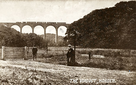 Postcard photograph entitled The Viaduct, Horden, showing the viaduct in the distance with a train crossing it; to the right of the photograph extensive woodland can be seen; in the foreground is a fence with a gate in it; in front of the fence two indistinct male figures are standing, one of whom appears to wearing a uniform