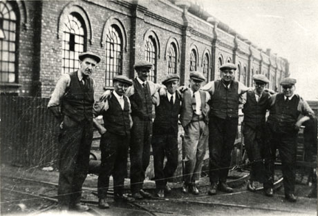Photograph of eight men standing in a line facing the camera with their arms on each others shoulders outside colliery buildings; the men are in shirt sleeves and have been identified as Officials at Horden Colliery during the 1926 Strike
