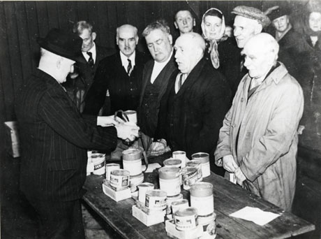 Photograph showing a man in a suit and Trilby hat standing at the left side of the photograph; he is photographed from the side and is standing behind a table on which piles of tinned food can be seen; in front of the table eleven people are standing; the photograph has been described as Sam Watson, Durham Miners' Association Secretary, issuing food parcels, Horden, 1939-1945