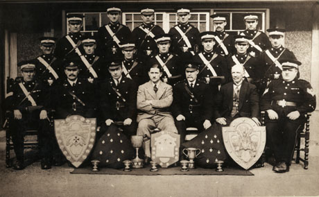 Photograph of eighteen men in uniform and two men in suits posed outside a building; in front of the men are ten trophies and two cushions on which badges appear to be pinned; the men have been described as members of the St. John Ambulance Brigade, Horden