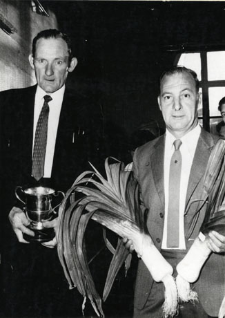 Photograph of a man holding a trophy cup and of a second man holding two large, presumably prize, leeks, which, also presumably, have won the cup held by the first man; the photograph has been described as Pop Porter With Leeks at Horden