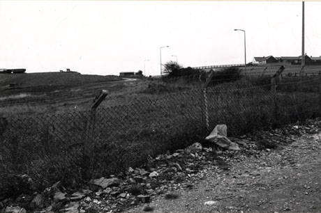 Photograph, described as Horden 1976, showing a wire fence across the picture from the bottom left-hand corner to the middle of the right of the picture; in front of the fence is the rough surface of an track and behind the fence rough grass; in the distance a low building on the brow of a hill at the right of the picture may be seen; high sodium street lights can be seen in the distance in the middle of the picture