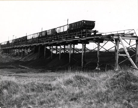 Photograph of coal trucks passing over a track built above coal waste and rough grass; the photograph is described as Coal Trucks Horden 1976; duplicate of hord 0112