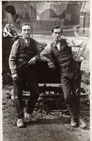 Photograph of two men dressed in shirt sleeves, waistcoats, and muddy shoes, and smoking cigarettes,leaning on a truck; in the background the end of a church building and houses can be seen indistinctly; the photograph has been described as Mr. Lowery and Mr. Mead Clearing The Dene at Blackhills Terrace, Horden, 1926