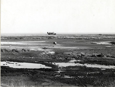 Photograph of the beach at Horden looking towards the sea; a lorry is parked in the distance near the sea; in the foreground the surface of the beach, covered in coal debris, pools of water and an empty oil drum, can be seen